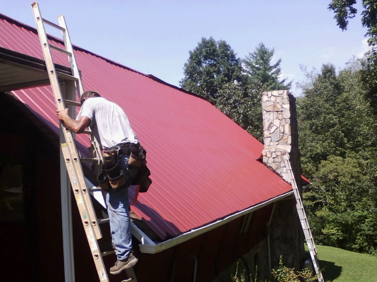 Roofer on a ladder beside a red metal roof and stone chimney — Soffit & Fascia Repair by Roofing Brothers in Waccabuc